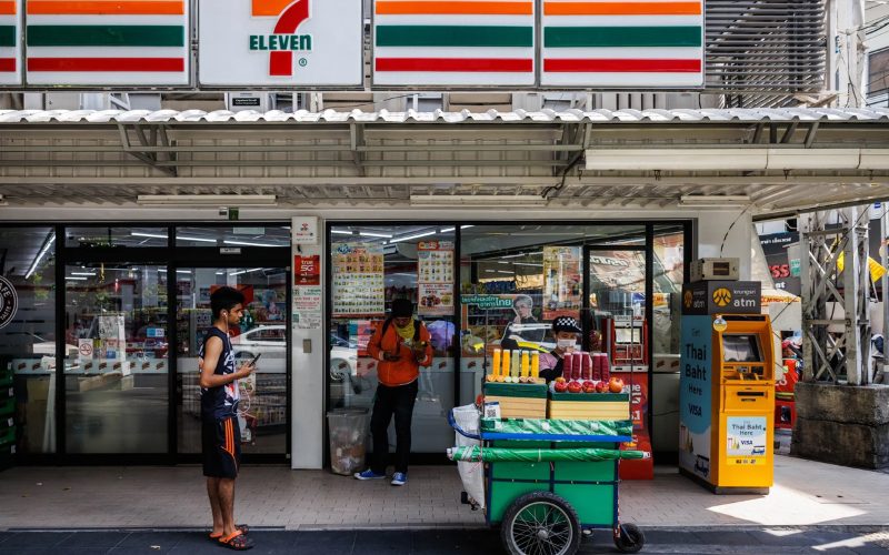 Façade d’un magasin 7-Eleven en Thaïlande, avec un distributeur automatique jaune Krungsri ATM et un vendeur ambulant proposant des jus de fruits frais devant l’entrée
