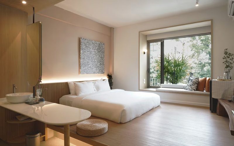 Chambre d’hôtel moderne et lumineuse en Thaïlande, décorée dans un style minimaliste avec un grand lit, des tons bois clairs et une baie vitrée donnant sur un jardin verdoyant