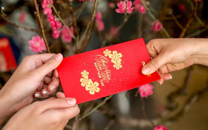 Deux personnes échangent une enveloppe rouge contenant de l’argent porte-bonheur (lì xì) devant des fleurs de pêcher, symbole traditionnel du Nouvel An lunaire vietnamien