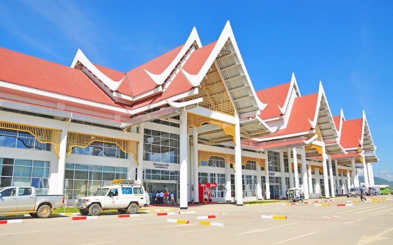 Façade de l’aéroport international de Luang Prabang, sous un ciel bleu clair, avec son architecture traditionnelle laotienne caractérisée par des toits rouges en pointe et des éléments décoratifs dorés