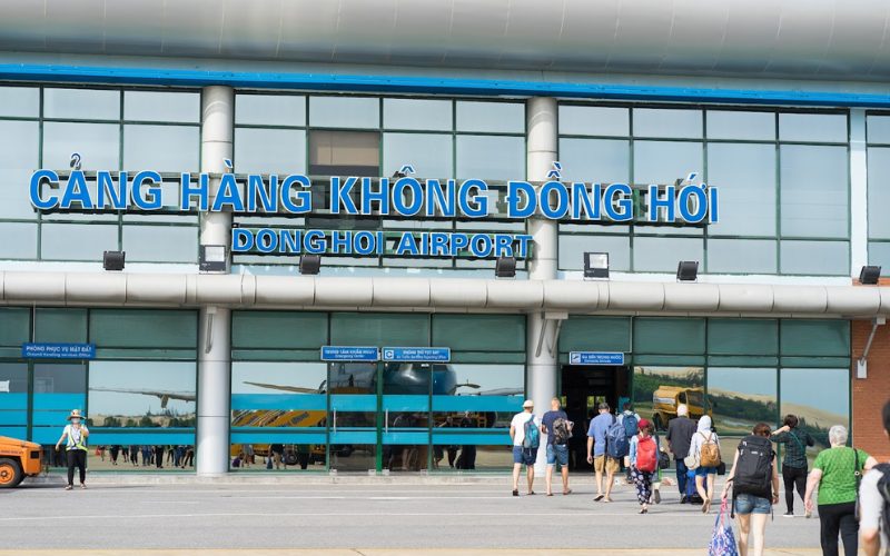 Façade de l'aéroport de Đồng Hới au Vietnam, avec des voyageurs entrant dans le terminal sous l'enseigne "Cảng Hàng Không Đồng Hới - Dong Hoi Airport"
