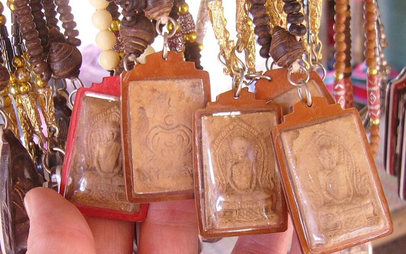 Amulettes bouddhistes thaïlandaises protégées dans des cadres en plastique, suspendues à des colliers de perles en bois, exposées dans un marché artisanal