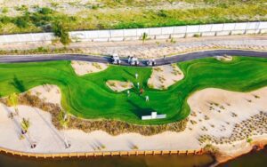 Vue aérienne d’un trou de golf bordé de sable avec des golfeurs en action, entouré de dunes et d’une route pour voiturettes
