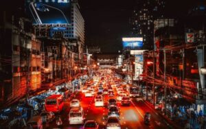 Scène nocturne animée à Bangkok, avec une avenue urbaine très fréquentée, des files de voitures aux feux rouges éclatants, des immeubles illuminés, des enseignes lumineuses et une atmosphère vibrante typique de la capitale thaïlandaise.