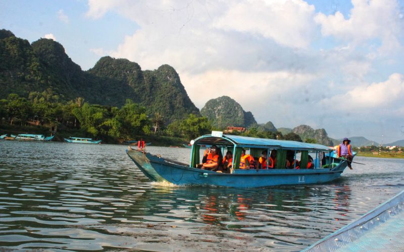 Bateau touristique transportant des passagers avec gilets de sauvetage sur la rivière de Phong Nha, entourée de montagnes karstiques verdoyantes sous un ciel partiellement nuageux