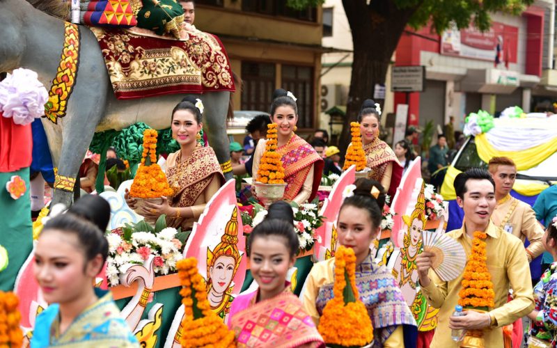Défilé coloré du Boun Pi Mai au Laos, avec des participants en tenues traditionnelles portant des offrandes de fleurs de souci, entourés de chars décorés et d’illustrations sacrées