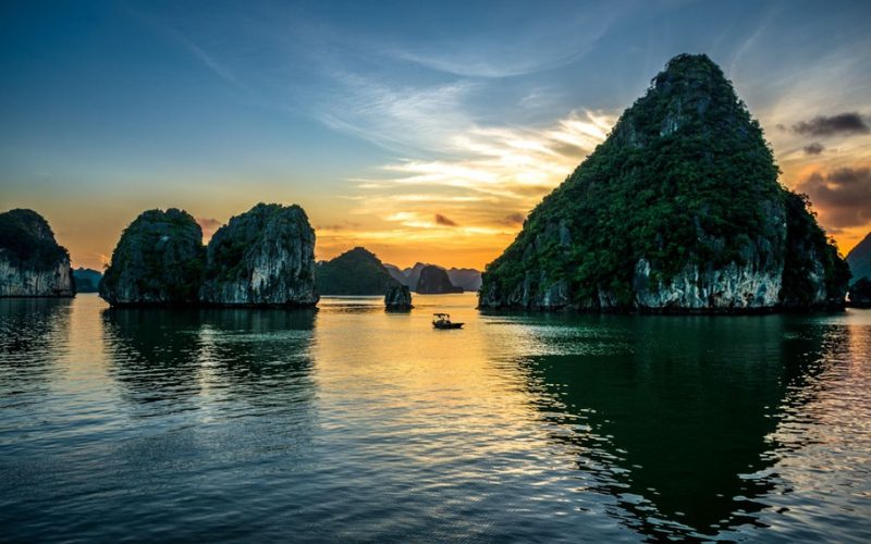 Baie d’Ha Long au coucher du soleil, avec des îlots karstiques verdoyants se reflétant sur une mer calme et un bateau traditionnel au centre