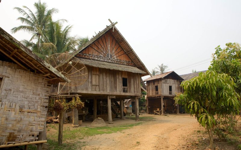 Maisons traditionnelles en bois sur pilotis dans un village rural, entourées de palmiers et de végétation tropicale