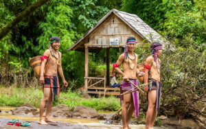 Trois hommes vêtus de costumes traditionnels ethniques debout devant une petite cabane en bois, entourés d'une végétation tropicale luxuriante. Ils sourient et tiennent des arcs et des paniers, illustrant une scène culturelle en plein air