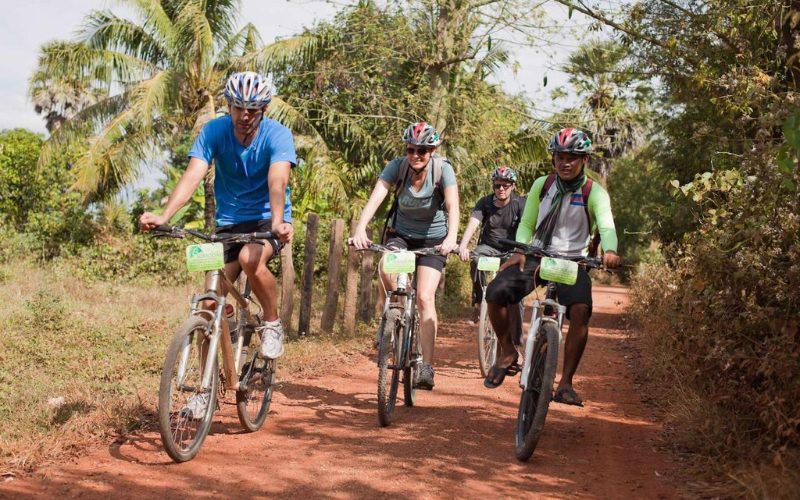 Un groupe de cyclistes explore la campagne de Battambang sur un chemin de terre rouge, entouré de palmiers et de végétation tropicale