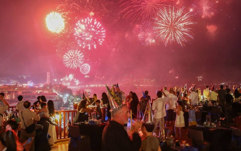 Invités réunis sur une terrasse pour une fête du Nouvel An, observant un grand feu d’artifice rouge illuminant le ciel et la baie, dans une ambiance festive et conviviale