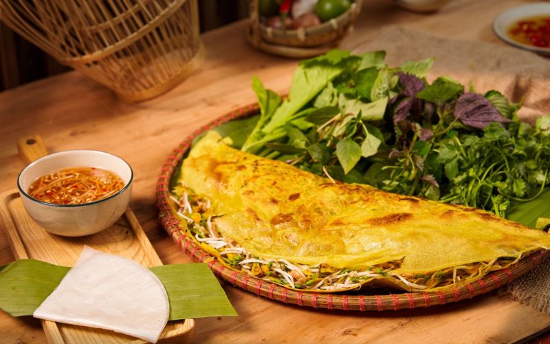 Un grand bánh xèo vietnamien croustillant, garni de germes de soja, crevettes et porc, servi avec un assortiment d’herbes fraîches, de galettes de riz et une sauce nuoc-mâm aigre-douce pour tremper