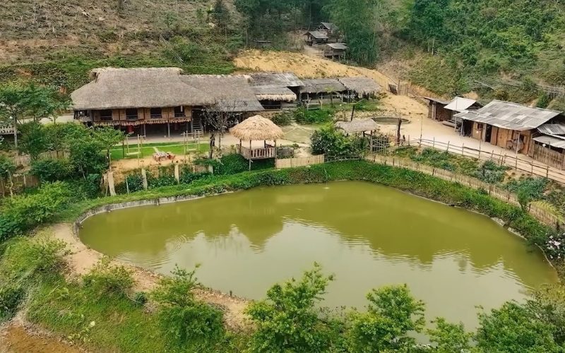 Vue d’ensemble d’un village traditionnel de Ban Lien, avec maison sur pilotis au toit de chaume, petit étang paisible et collines verdoyantes en arrière-plan.