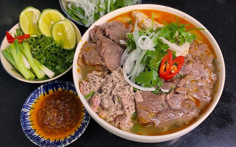Bol de bún bò Huế, une soupe vietnamienne épicée à base de nouilles de riz, de bœuf, d’herbes fraîches, de piments rouges, d’oignons émincés et de coriandre, accompagné de citron vert