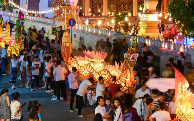 Foule de personnes célébrant le festival bouddhiste Boun Ok Phansa, entourée de lanternes illuminées en forme de bateaux-dragons et de décorations colorées, dans une ambiance festive et spirituelle en soirée