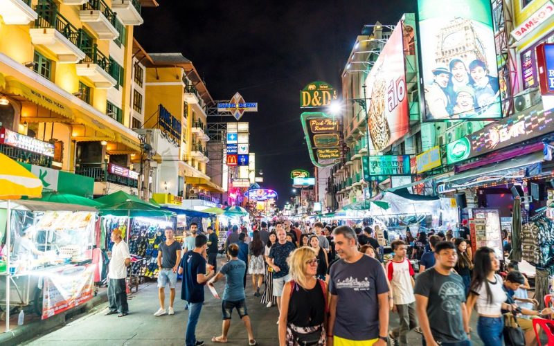 Rue animée de Khao San Road à Bangkok de nuit, illuminée par des enseignes lumineuses et bordée d’étals de street food, de bars et de boutiques, remplie de touristes et locaux flânant dans une ambiance festive