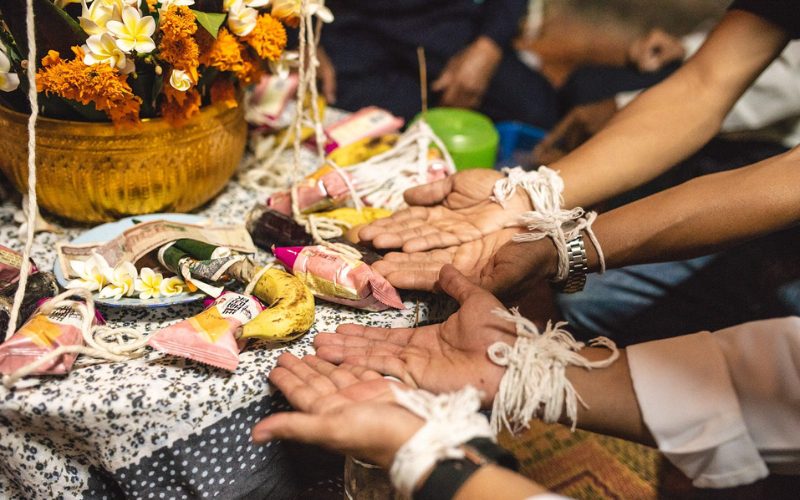 Cérémonie du Baci au Laos, avec des poignets noués de fils sacrés et des offrandes symboliques, dont des fleurs, des fruits et des billets de banque