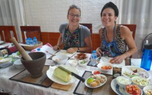 Deux voyageuses souriantes participent à un cours de cuisine traditionnelle laotienne, assises à une table garnie d’ingrédients frais, d’épices et d’ustensiles locaux, prêtes à préparer des plats typiques du Laos