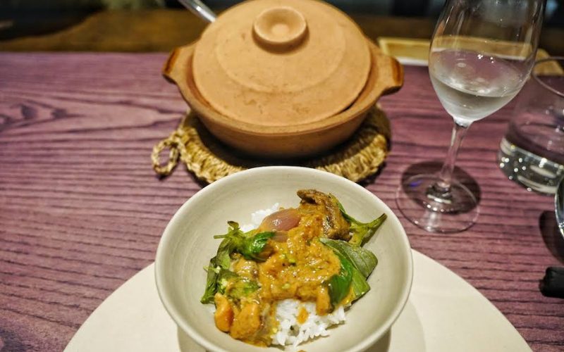 Bol de riz garni d’un curry vietnamien aux légumes et herbes fraîches, posé sur une assiette blanche, accompagné à l’arrière d’un pot en terre cuite et d’un verre d’eau sur une table en bois mauve élégante
