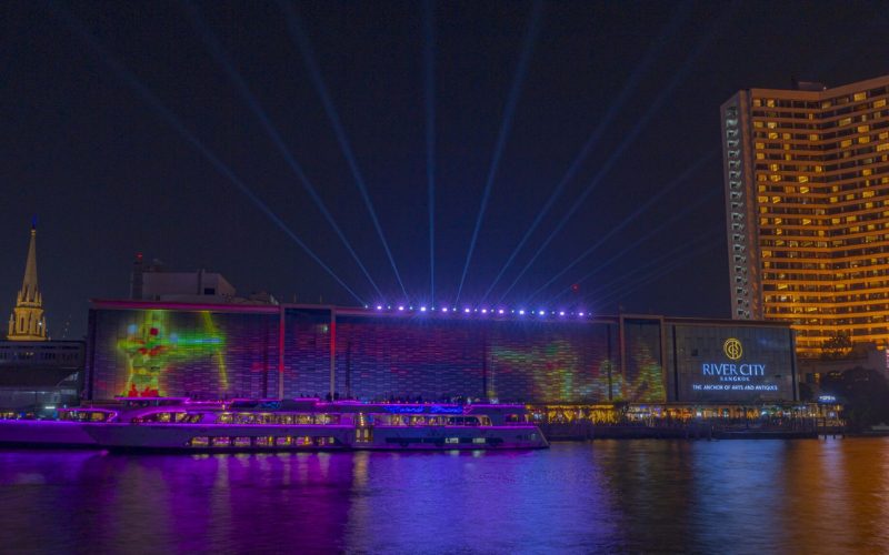 Façade de River City Bangkok illuminée par un spectacle de projections colorées et de faisceaux lumineux, avec des bateaux de croisière éclairés naviguant sur la rivière Chao Phraya la nuit