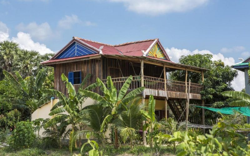 Champa Lodge, entourée de bananiers et de verdure tropicale, avec un toit rouge, une décoration colorée en façade et un balcon en bois offrant une vue sur la campagne environnante