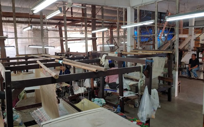 Atelier de tissage traditionnel avec plusieurs métiers à tisser en bois, où des artisans locaux fabriquent manuellement des étoffes, dans une ambiance artisanale et authentique au cœur d’un bâtiment simple mais animé