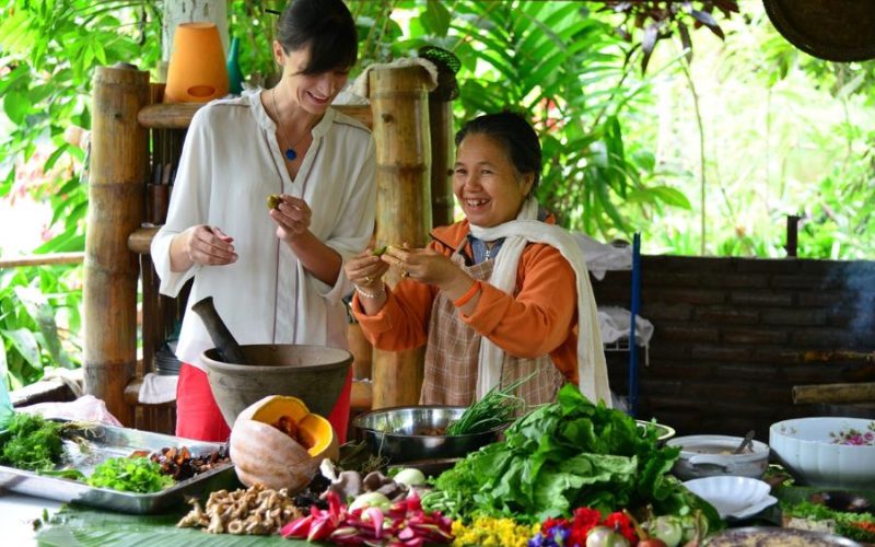 Une femme locale enseigne à une voyageuse comment cuisiner un plat traditionnel lao, entourées d'ingrédients frais et colorés dans une cuisine en plein air au cœur de la nature