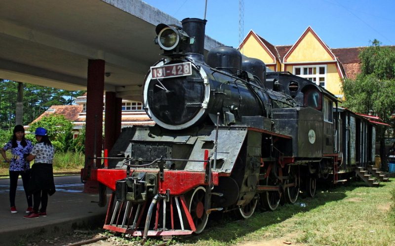Ancienne locomotive à vapeur exposée à la gare de Da Lat au Vietnam, avec des visiteurs en arrière-plan et un bâtiment de style colonial