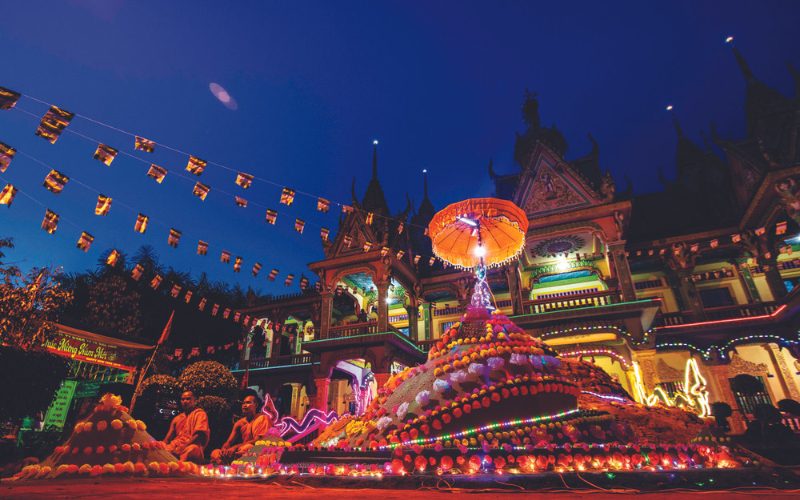 Monticules de sable décorés de lumières et d’offrandes devant une pagode khmère illuminée au crépuscule, tandis que des moines célèbrent le Nouvel An cambodgien, Chol Chnam Thmay