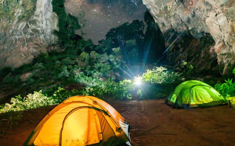 Tentes de camping éclairées installées à l'intérieur d'une grotte ouverte à Phong Nha, avec un ciel étoilé visible à travers l'ouverture et des parois rocheuses imposantes autour