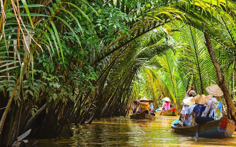 Touristes en barques traditionnelles naviguant sur un canal étroit bordé de palmiers d’eau à Vinh Long, au cœur de la végétation dense du delta du Mékong