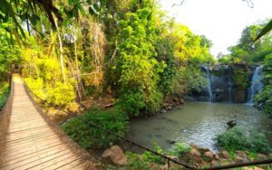Pont suspendu en bois traversant une forêt tropicale luxuriante, menant à une cascade entourée de rochers et de végétation verdoyante, baignée par la lumière dorée du soleil en fin de journée