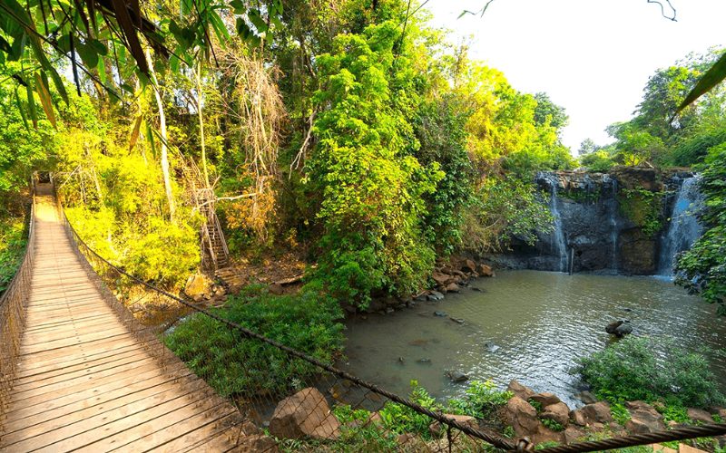 Pont suspendu en bois traversant une forêt tropicale luxuriante, menant à une cascade entourée de rochers et de végétation verdoyante, baignée par la lumière dorée du soleil en fin de journée