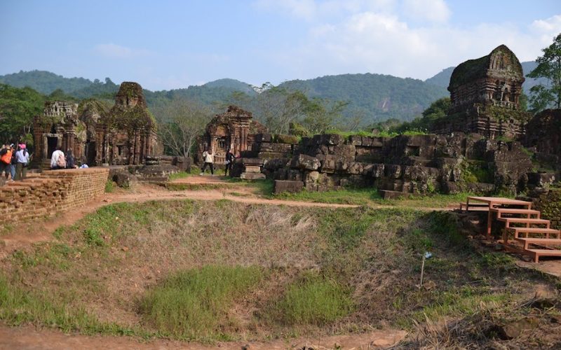 Cratère de bombe au milieu des ruines du sanctuaire de My Son, entouré de temples anciens en briques rouges, avec des visiteurs explorant le site sous un ciel bleu clair