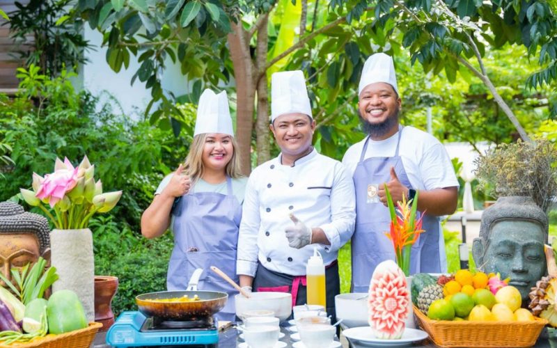 Trois participants souriants en tenue de chef lors d’un atelier de cuisine khmère en plein air, entourés de fruits tropicaux, de légumes frais et de décorations traditionnelles dans un cadre verdoyant