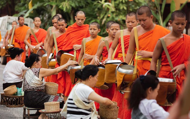 Cérémonie matinale d’aumônes à Luang Prabang: des moines bouddhistes en robe orange reçoivent des offrandes de nourriture de la part de fidèles assises au bord de la route, une tradition spirituelle emblématique du pays
