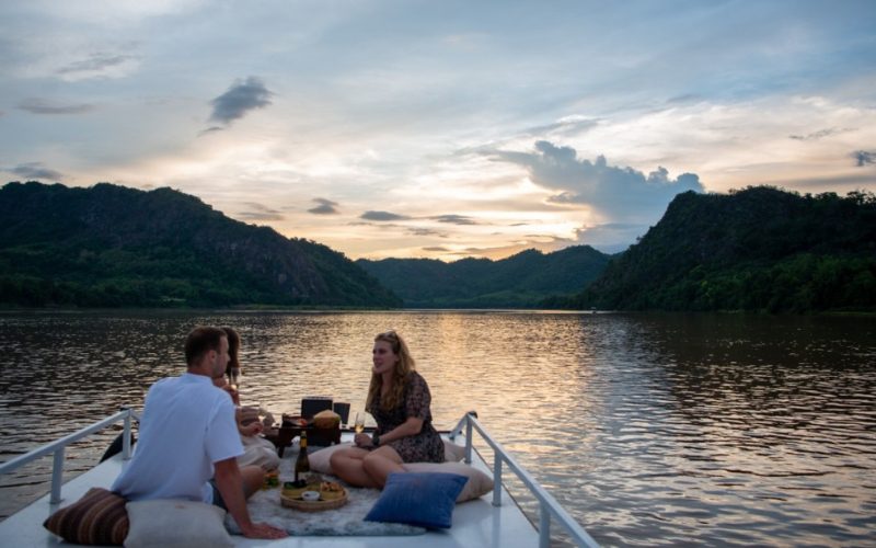 Couple profitant d’un dîner romantique sur un bateau au coucher du soleil sur le Mékong, entouré de montagnes verdoyantes au Laos