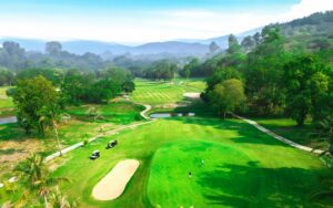 Vue aérienne d’un parcours de golf niché au cœur d’un paysage montagneux verdoyant, avec fairways bien entretenus, greens luxuriants, voiturettes de golf et golfeurs en pleine partie sous un ciel dégagé