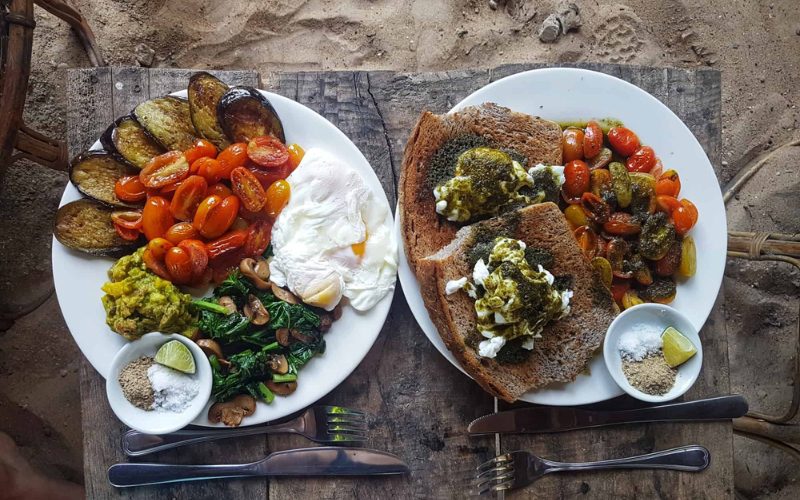 Assiettes colorées servies sur une table en bois sur le sable : légumes grillés, tomates cerises, œufs pochés, pain grillé et condiments pour un repas sur la plage