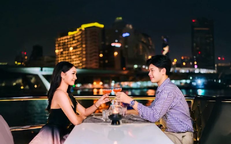 Couple en dîner romantique sur un bateau de croisière à Bangkok, trinquant au coucher de la nuit avec la skyline illuminée en arrière-plan