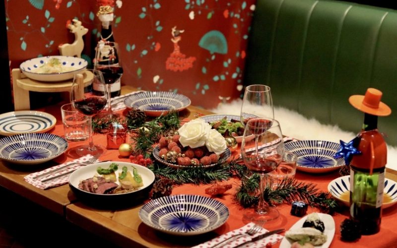 Table de fête décorée pour un dîner de Noël, avec vaisselle colorée, verres de vin rouge, bougies, branches de sapin et plats raffinés disposés dans une ambiance chaleureuse