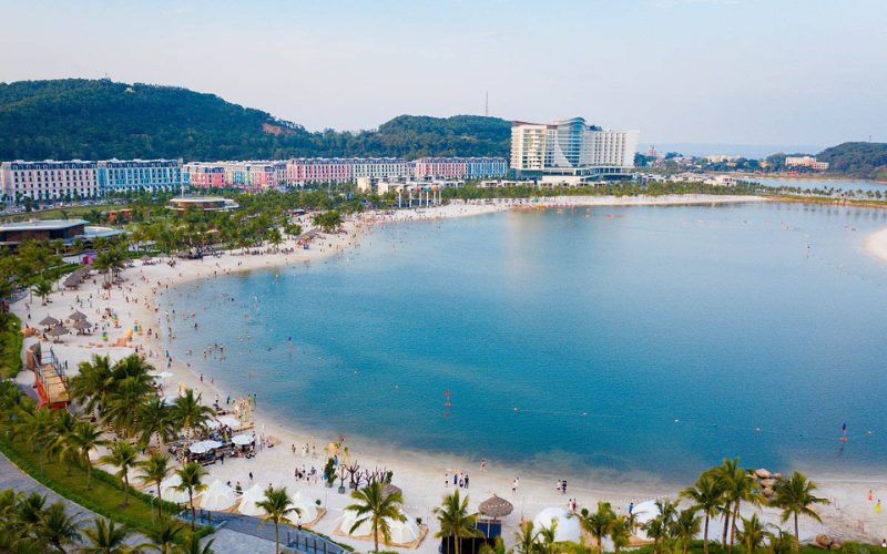 Vue panoramique de la plage artificielle de Bai Chay à Ha Long, bordée de palmiers, d’hôtels modernes et de bâtiments colorés. De nombreux visiteurs profitent du sable blanc et des eaux calmes, créant une atmosphère animée et estivale au bord de la baie