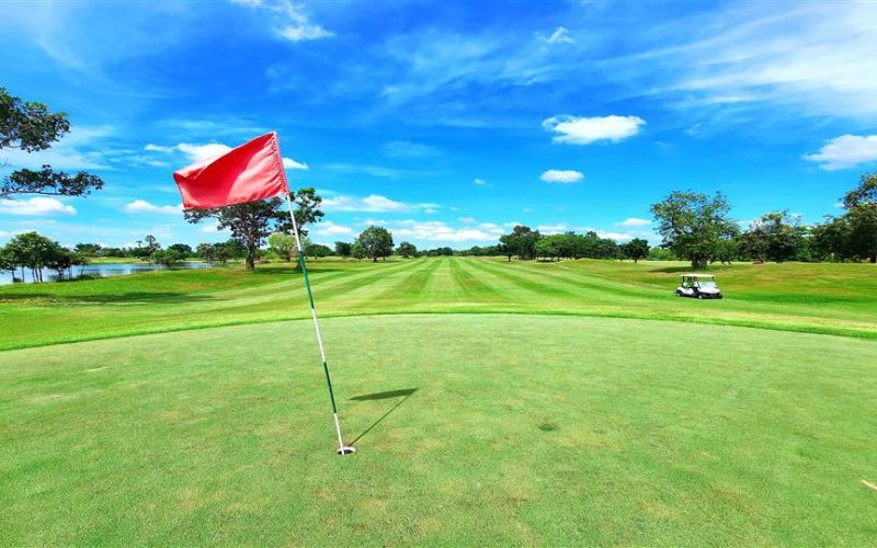 Vue d’un green de golf avec un drapeau rouge en premier plan, donnant sur un fairway parfaitement entretenu sous un ciel bleu éclatant, avec une voiturette de golf visible à l’arrière-plan