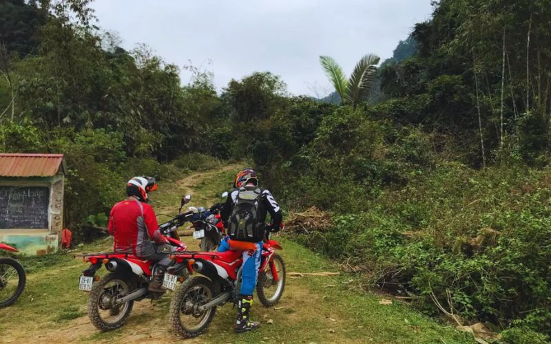 Deux motards en tenue de protection s’apprêtent à s’aventurer sur un sentier de terre traversant la jungle, au départ d’un petit village en pleine nature
