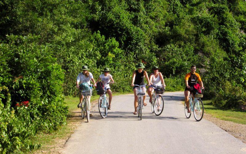 Groupe de touristes à vélo sur une route de campagne entourée de végétation luxuriante, sous un ciel ensoleillé, dans un cadre naturel paisible au Vietnam