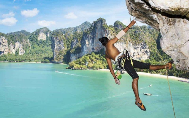 Grimpeur pratiquant l’escalade sur une falaise surplombant la mer d’Andaman à Railay Beach, Krabi, avec une vue panoramique sur les falaises et les eaux turquoise