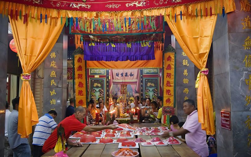 Célébration du festival Por Tor dans un temple chinois décoré de rideaux et bannières colorés, avec des fidèles présentant des offrandes de gâteaux rouges en forme de tortue sur une grande table en bois