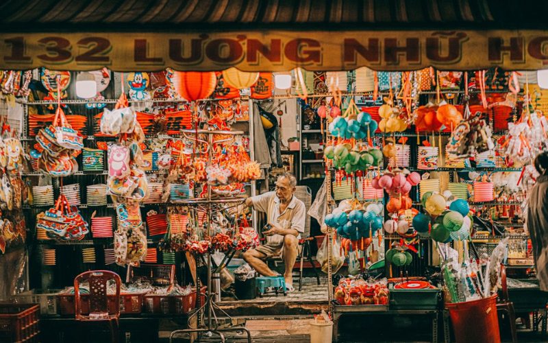 Un vendeur âgé assis dans une boutique traditionnelle de lanternes, entouré de décorations colorées du Tết Trung Thu, notamment des lampions en papier