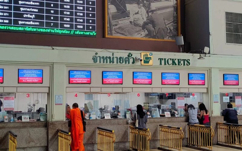 Comptoirs de billetterie de la gare de Bangkok avec des voyageurs, dont un moine bouddhiste, achetant leurs billets de train sous les panneaux d’affichage électroniques