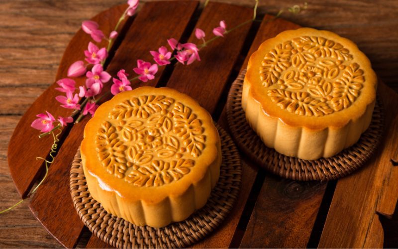 Deux gâteaux de lune traditionnels vietnamiens (bánh nướng) joliment décorés, posés sur des dessous en rotin, accompagnés de fleurs roses sur une planche en bois rustique
